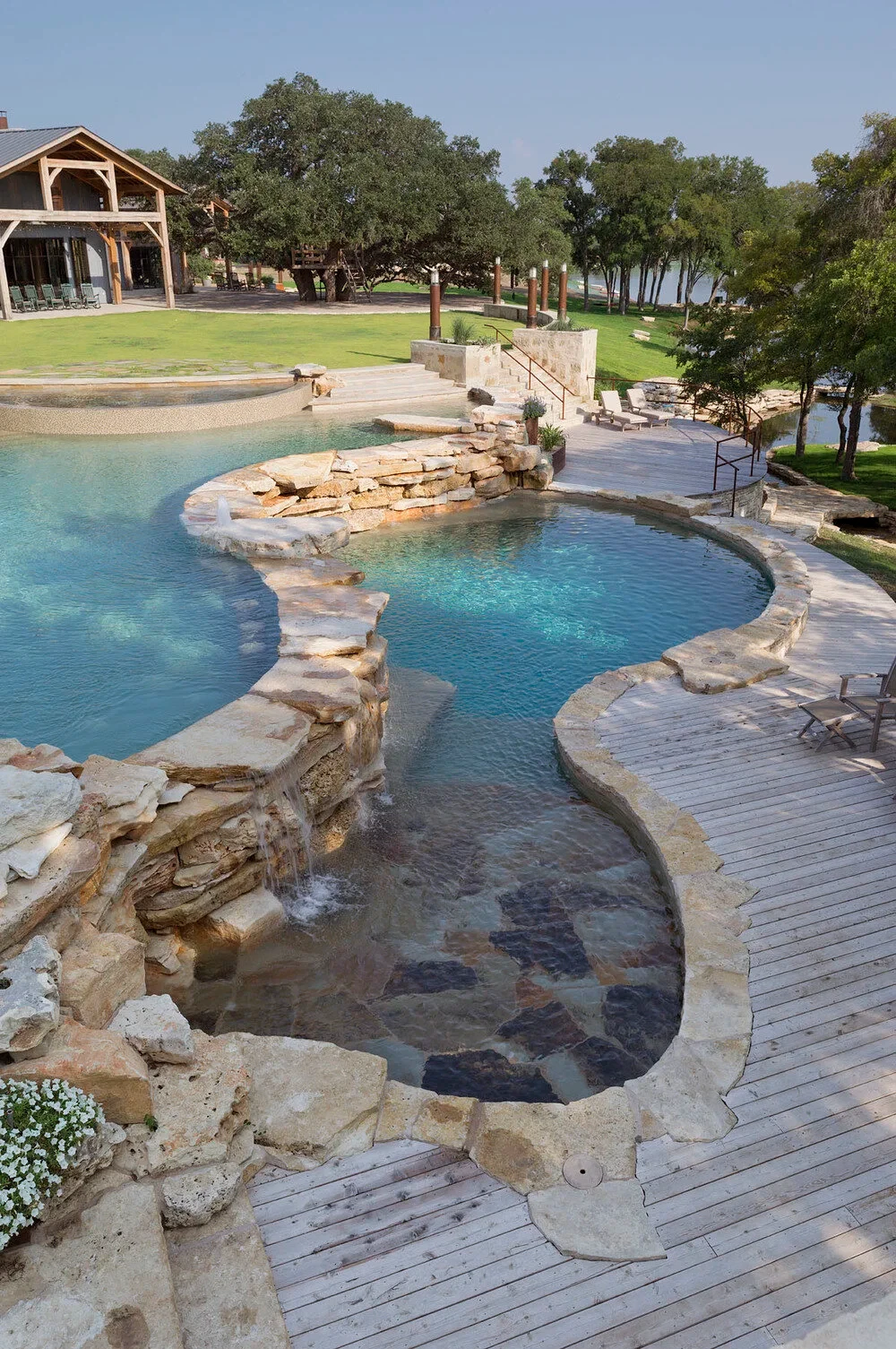 Freeform pool with a flagstone waterfall, shallow ledge and wood deck beside a lakeside lodge and oaks