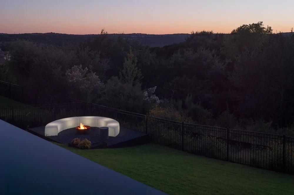 Curved glowing fire pit sofa on a lawn above the Hill Country at dusk, beside an infinity pool