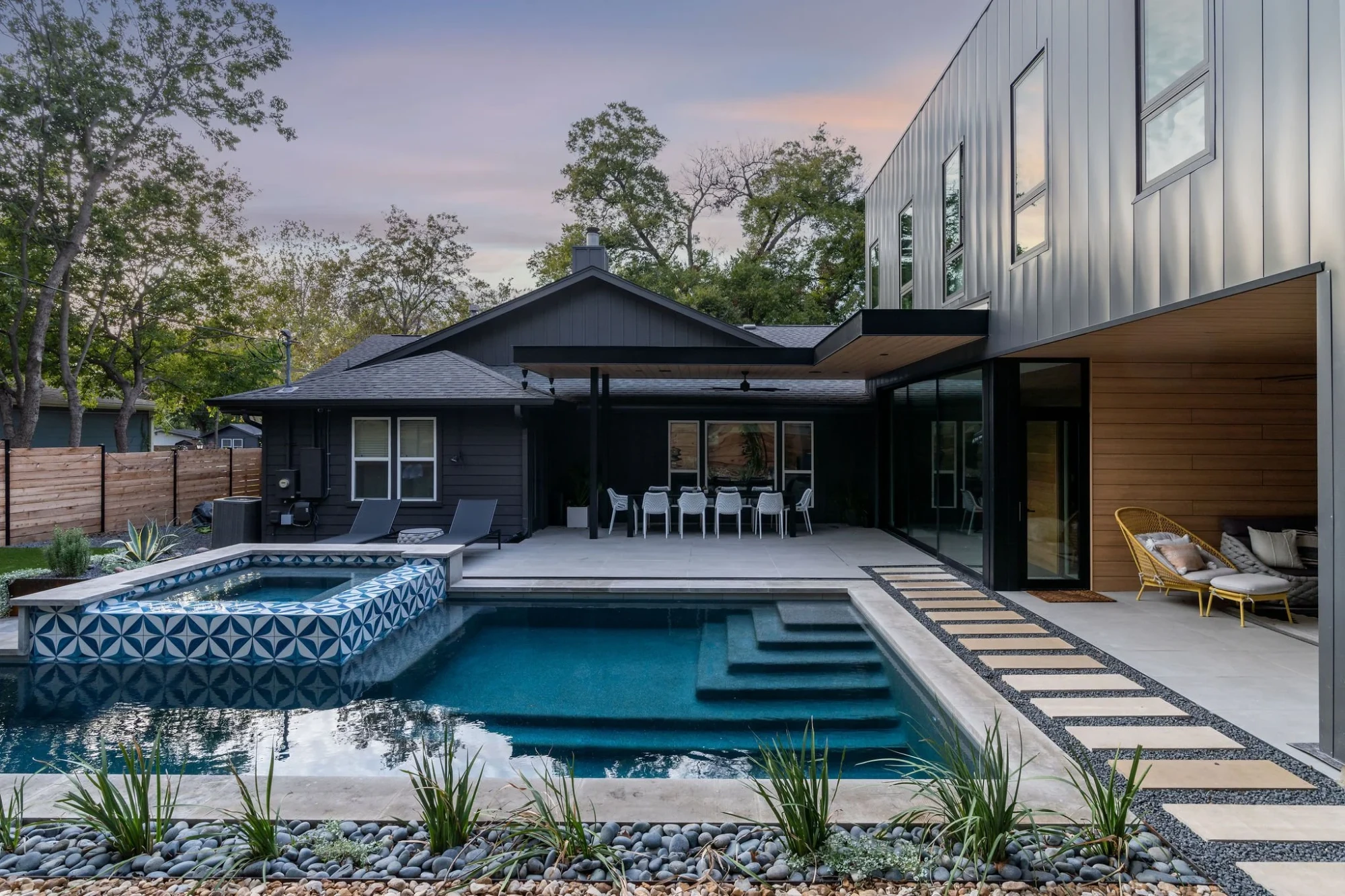 Geometric pool with blue-tile spa and steps beside a black modern home and patio at dusk in Austin