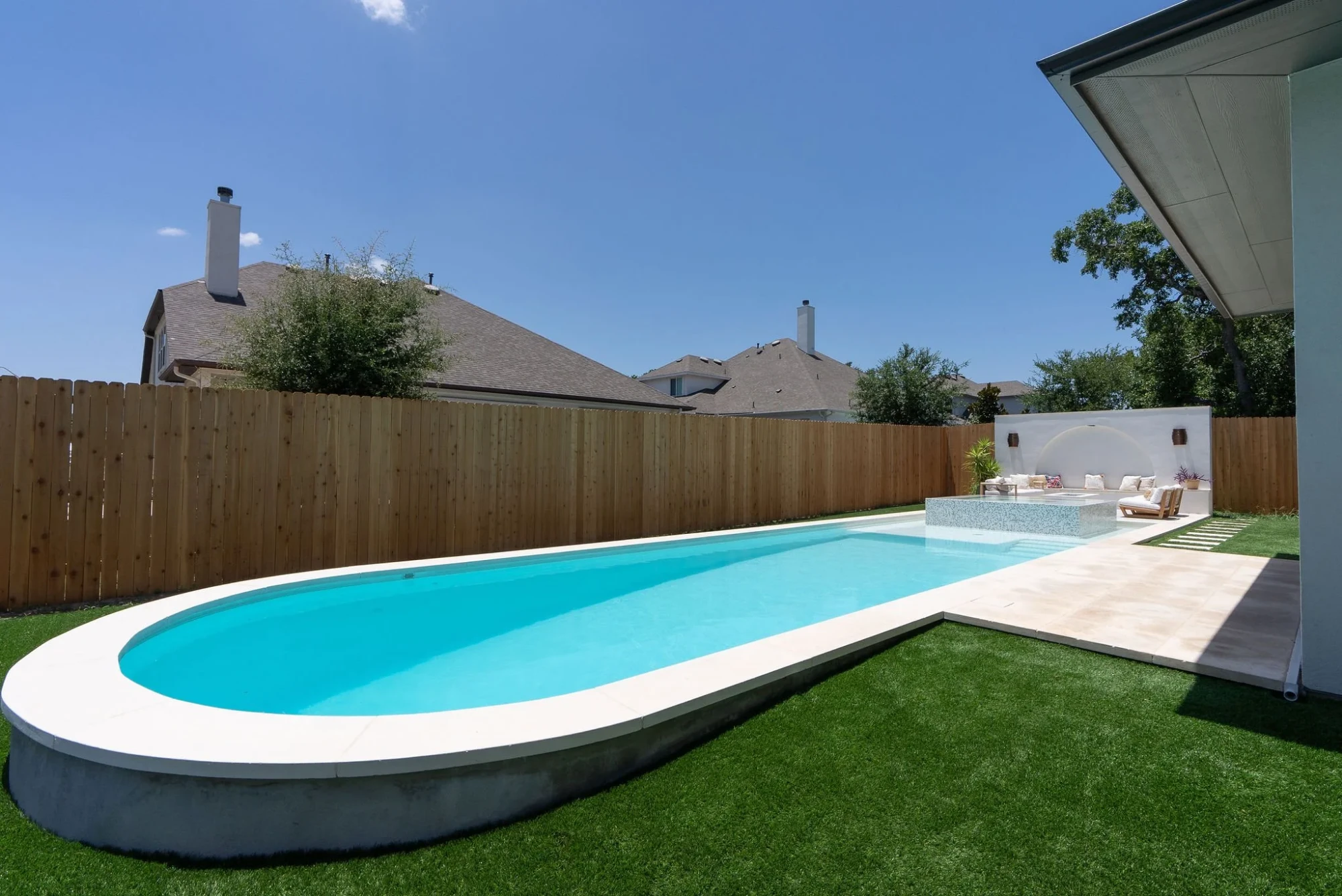 Long geometric pool with a rounded end and white coping on artificial turf, bordered by a wood fence in Austin