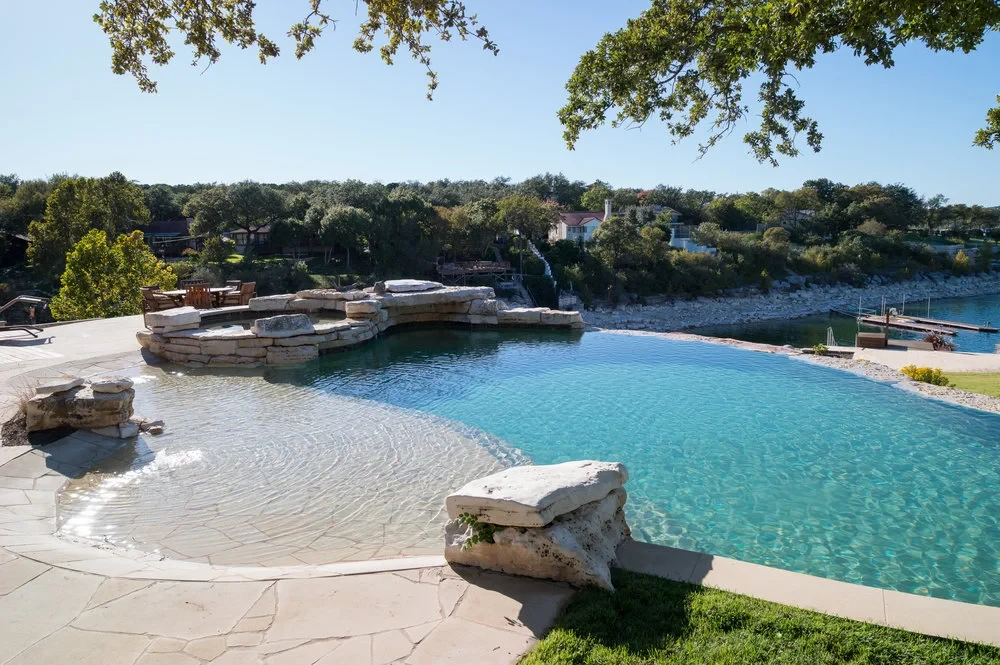 Curving infinity pool over Lake Travis with a raised stone spa and boulder accents on a flagstone deck