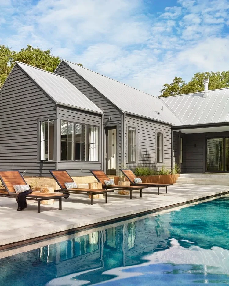 Gray board-and-batten home with a metal roof beside a rectangular pool and teak lounge chairs