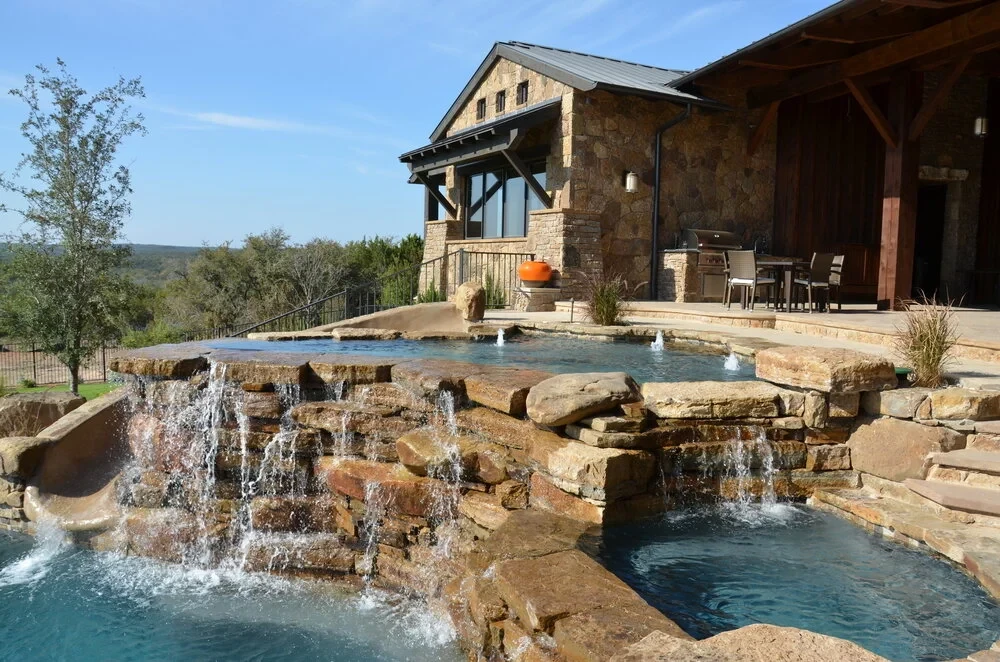 Freeform pool spilling over layered stone waterfalls below a stone home with a covered patio and Hill Country view