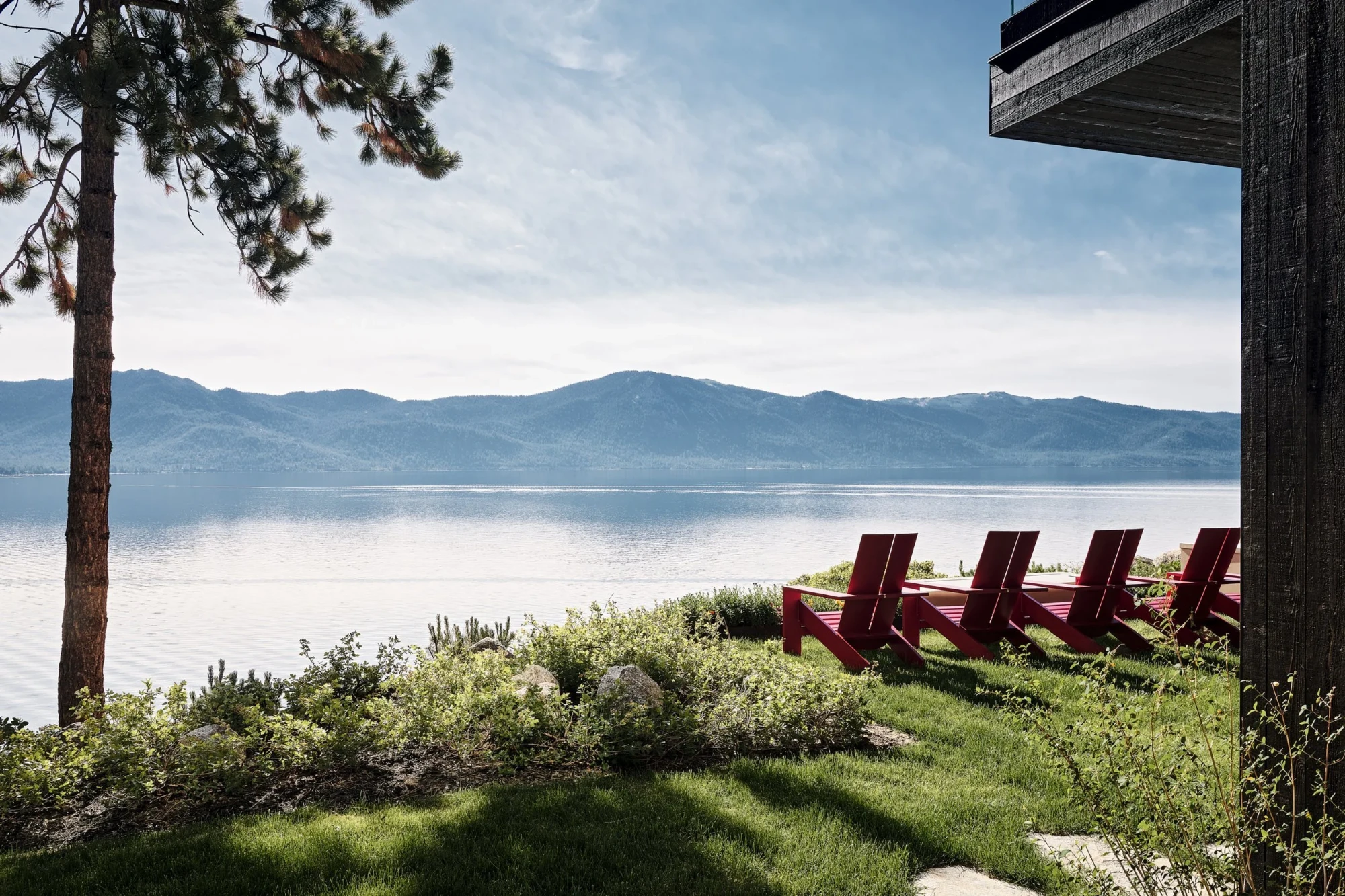 Red Adirondack chairs on a grassy bluff above a calm lake, framed by pines and distant mountains