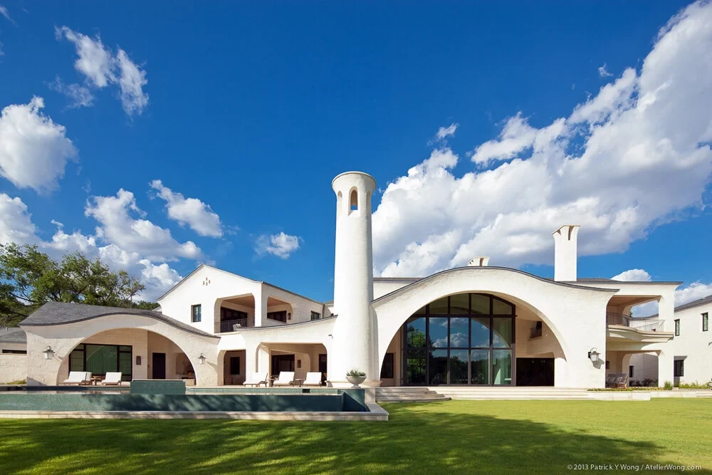 White-stone estate with sweeping curved arches and a round tower under big clouds, dark pool below
