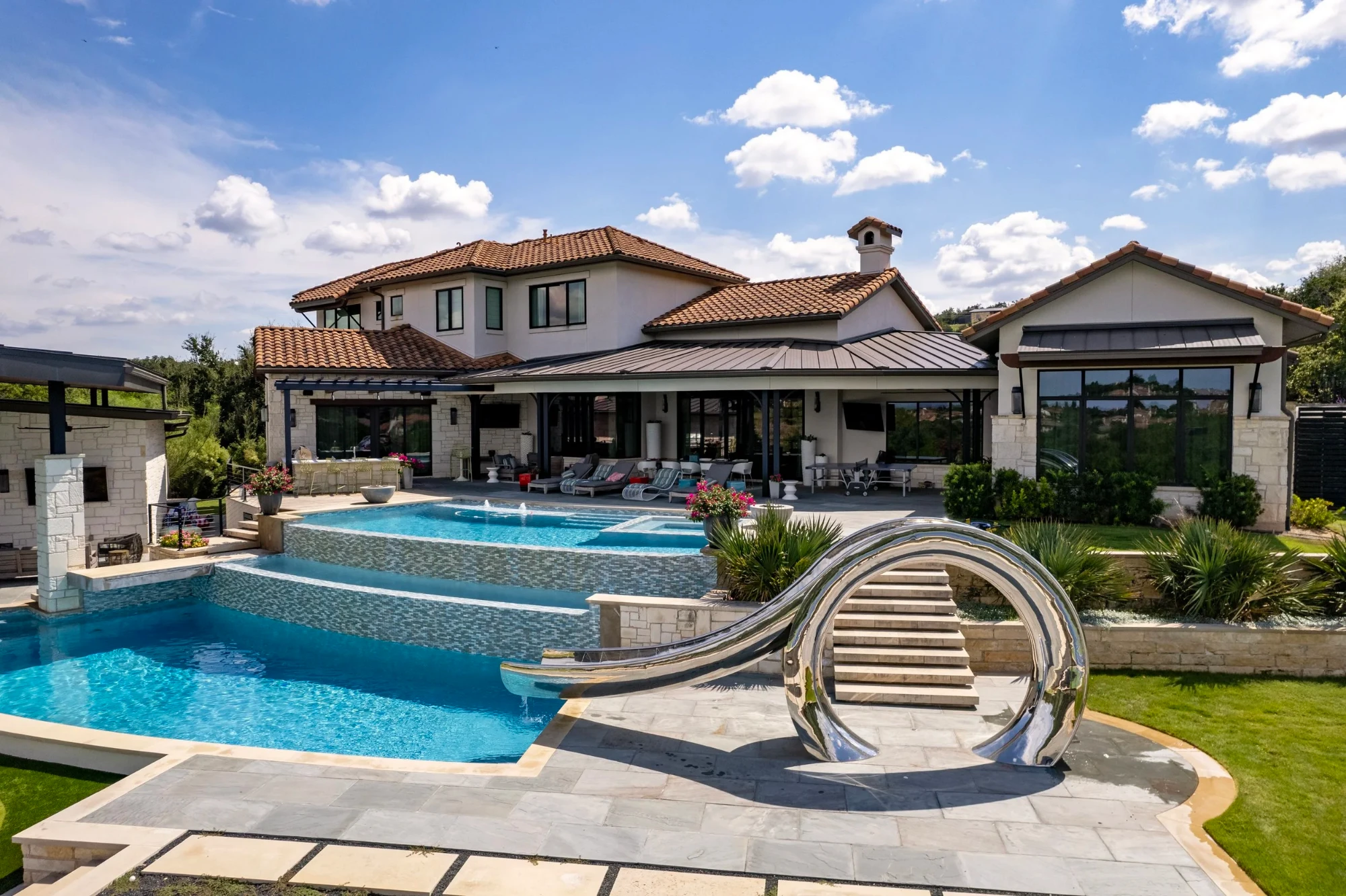 Stainless waterslide spilling into a blue-tile pool, with Lakeway hills and rooftops in the distance