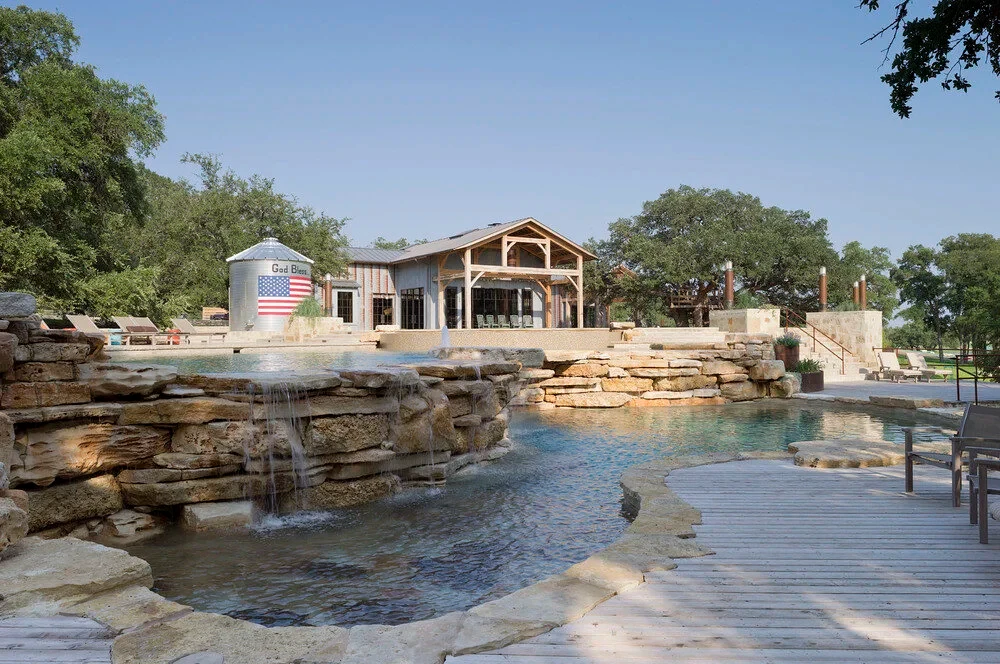 Freeform pool with stone waterfalls, a tanning ledge and wood deck below a timber lodge and lake