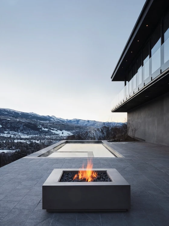 Square fire pit flaming on a stone terrace beside an infinity pool over the snowy Aspen mountains