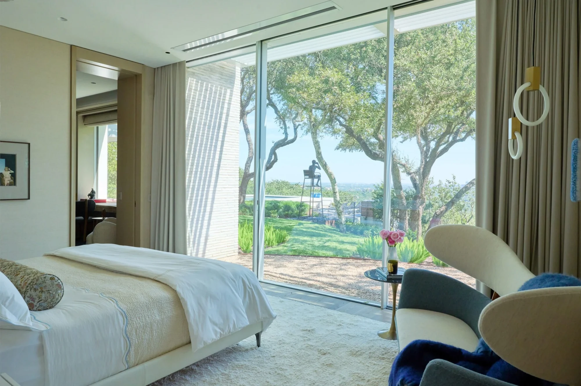 Serene bedroom with floor-to-ceiling glass framing live oaks and a Hill Country view, white bed and armchairs