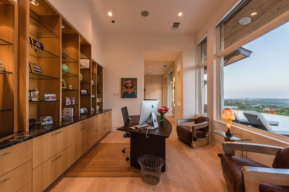 Home office with wood built-in shelves, a dark desk, and tall windows over the Hill Country at dusk