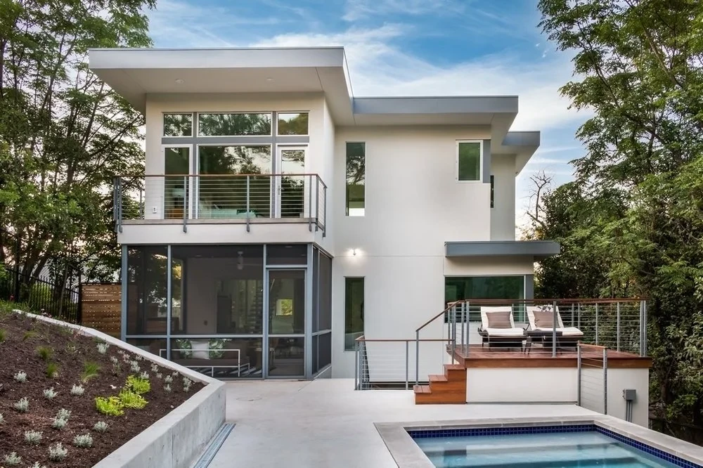 Modern white stucco home on a wooded hillside in Travis Heights, with a glass living room and pool below