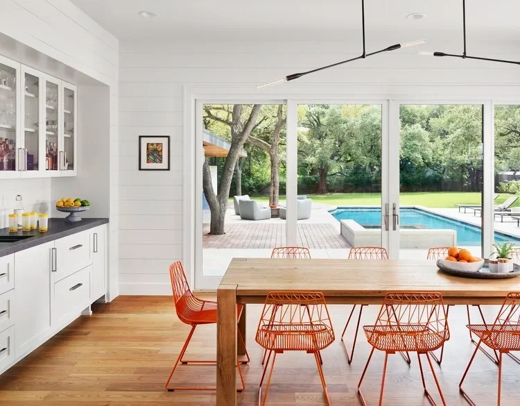 View from a white shiplap dining room through glass doors to a backyard pool, deck and live oaks