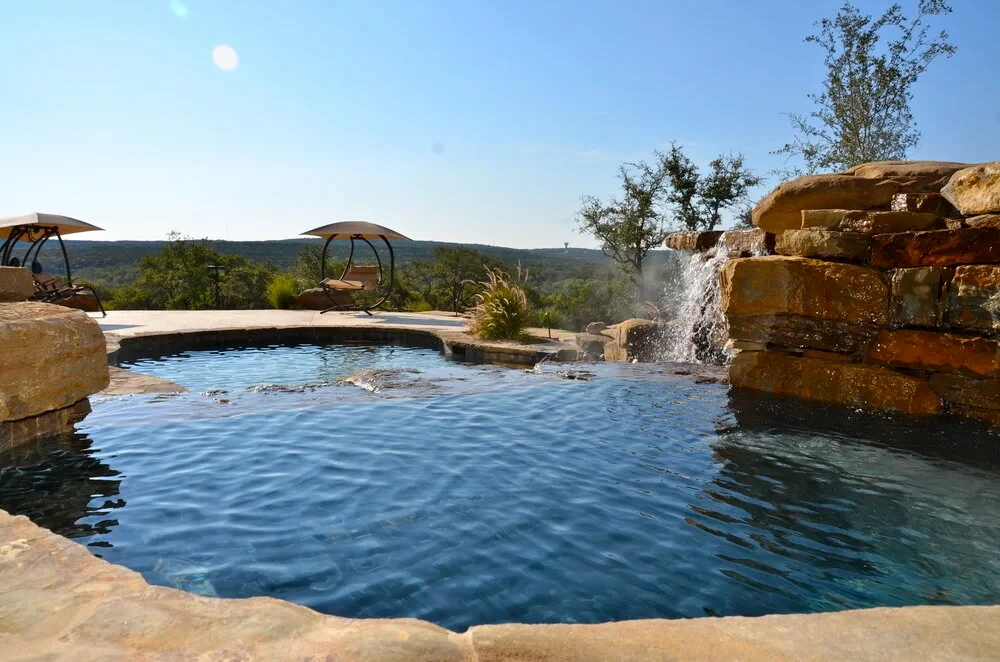 Misty freeform pool with stone-edged waterfalls, shade umbrellas and Hill Country views beyond the deck