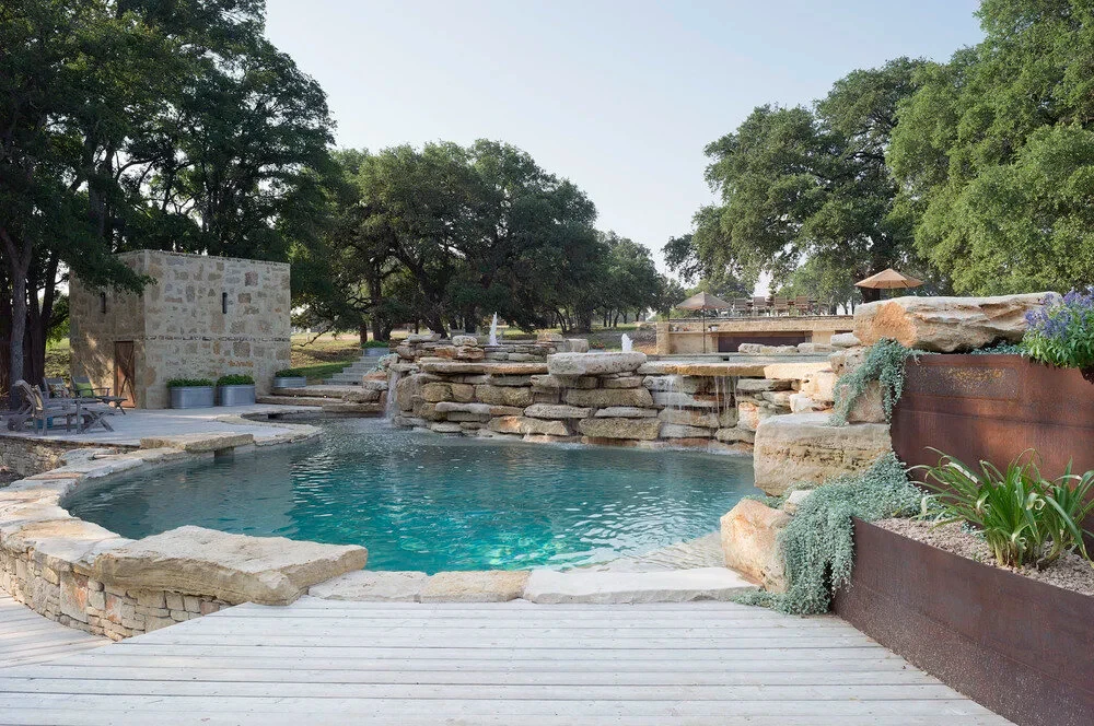 Rock waterfall and flagstone deck of a freeform pool, with a timber lodge beyond the oaks