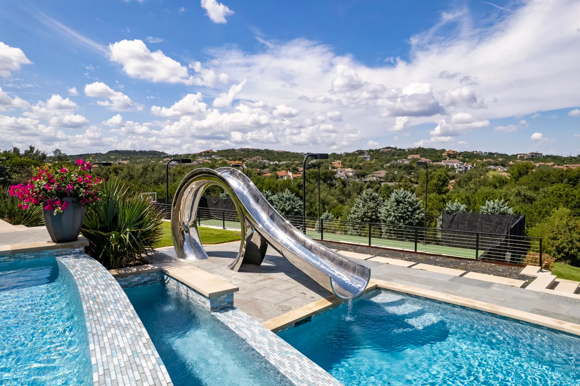Stainless steel waterslide pouring into a glass-tile pool above a sweeping Lakeway hillside view