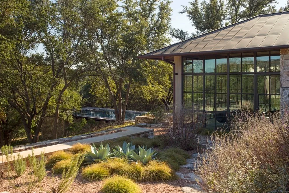 Glass-walled pavilion above a pool and native grasses on a wooded Hill Country slope at dusk