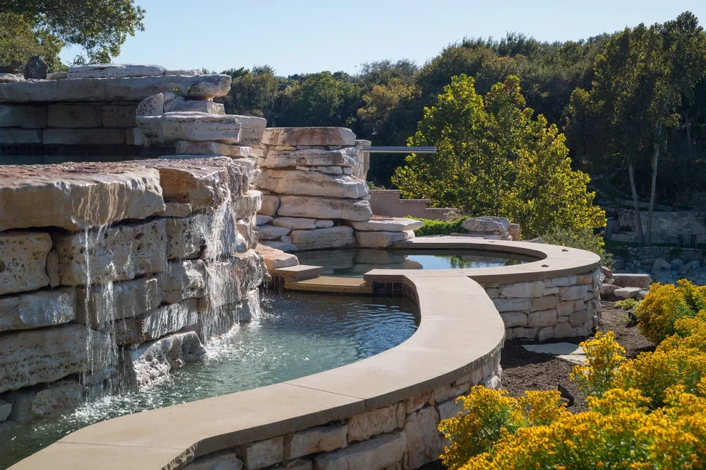 Stacked-stone waterfall spilling into a curved spa with stone coping and Hill Country trees beyond