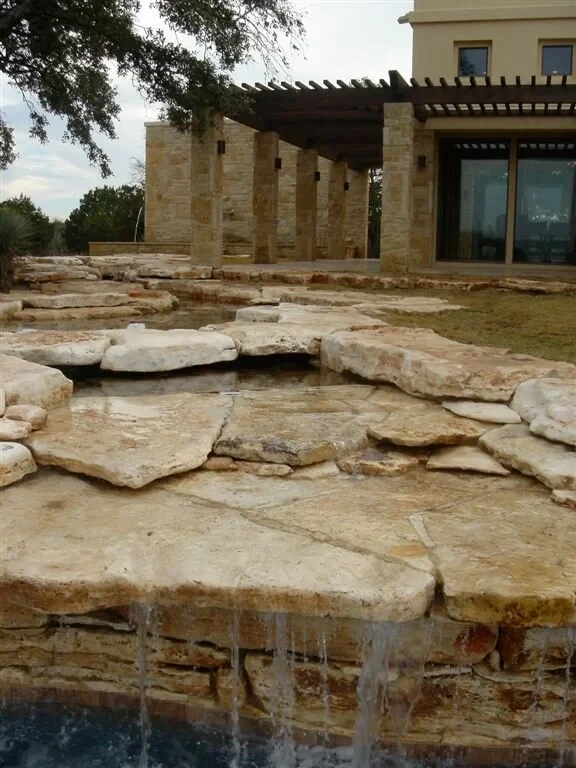 Limestone-slab waterfall spilling into a pool below a stone home with a pergola and glass doors