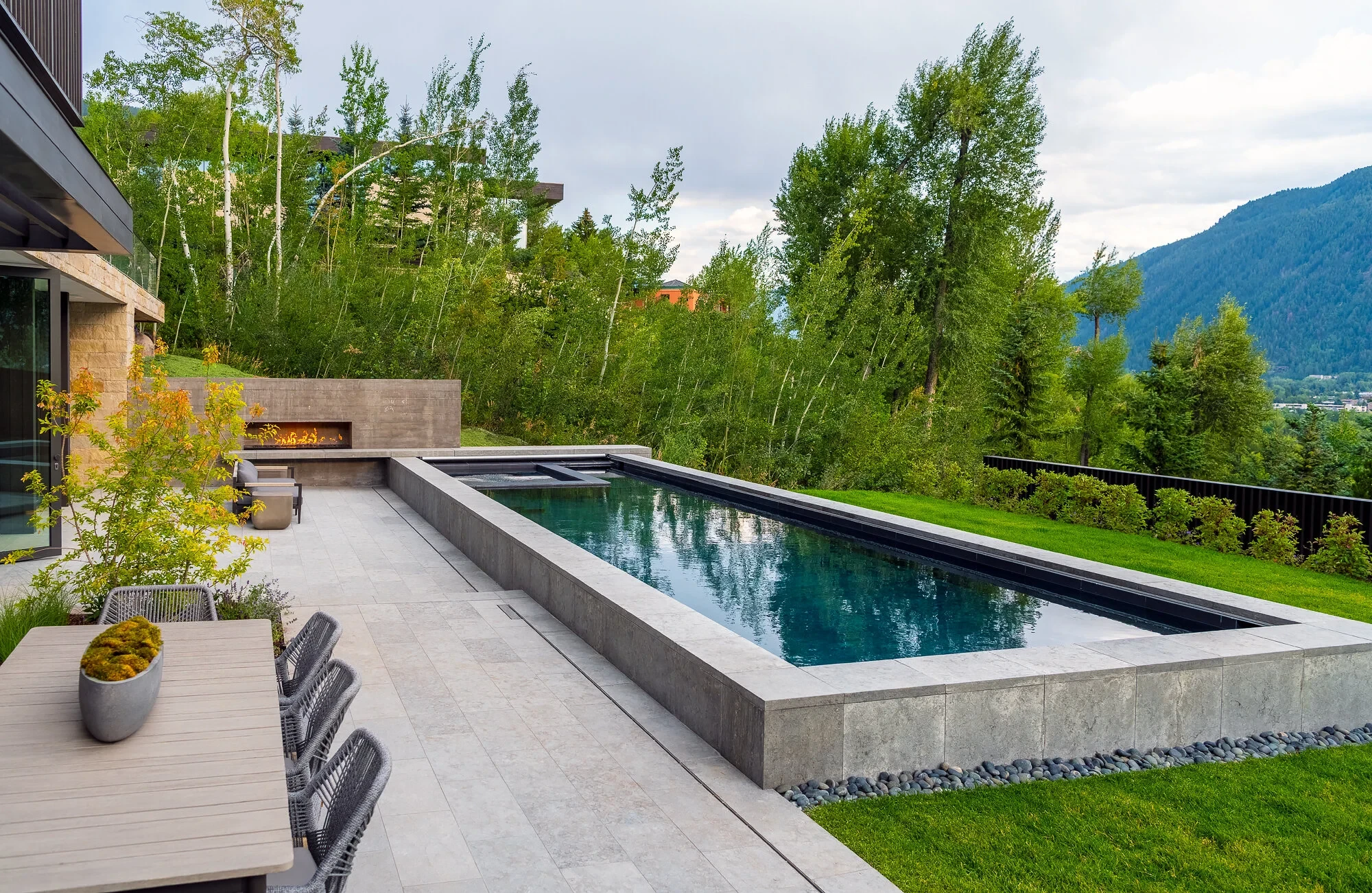 Long geometric pool with stone coping beside a fireplace and lawn, backed by aspens and mountains