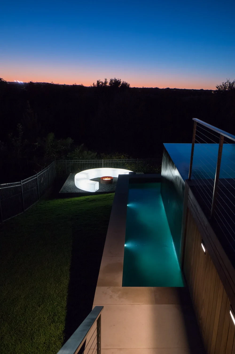 Lap pool and glowing circular fire pit lounge on a lawn above the Hill Country at twilight