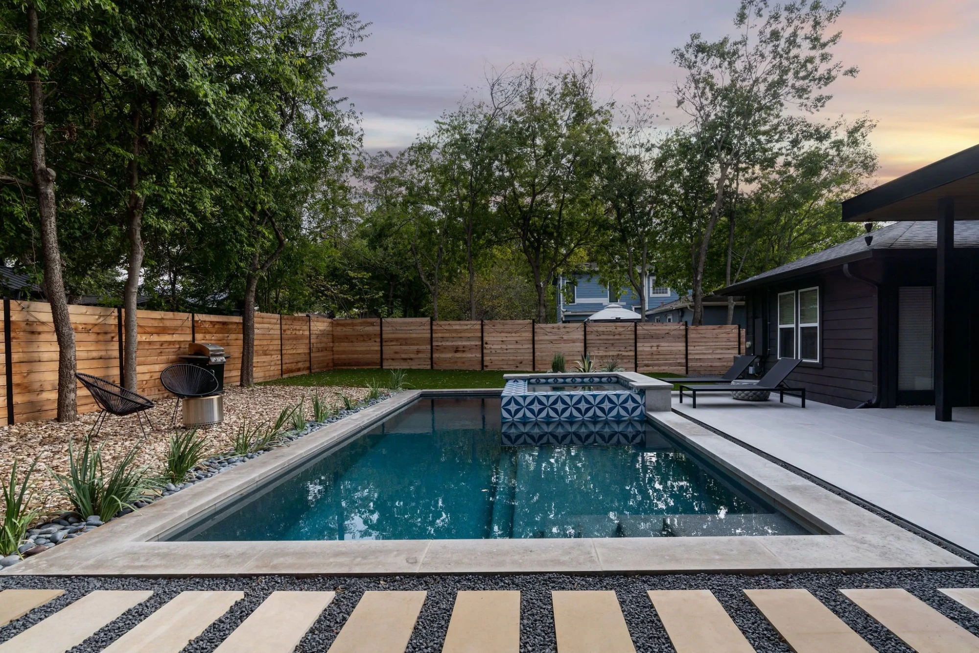 Geometric pool with a blue-tile spa in a fenced backyard with stone paths at dusk in Austin