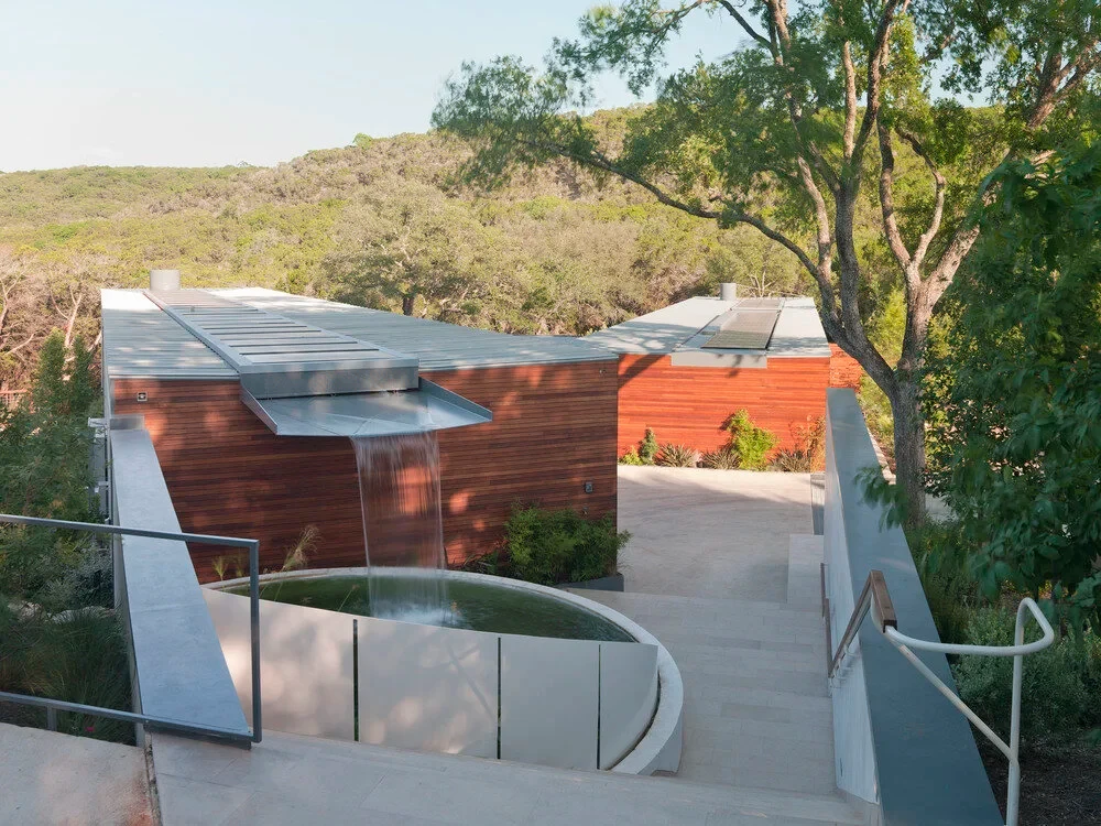 Wood-clad home with metal roofs on a hillside, curved glass-rail stairs above the Hill Country