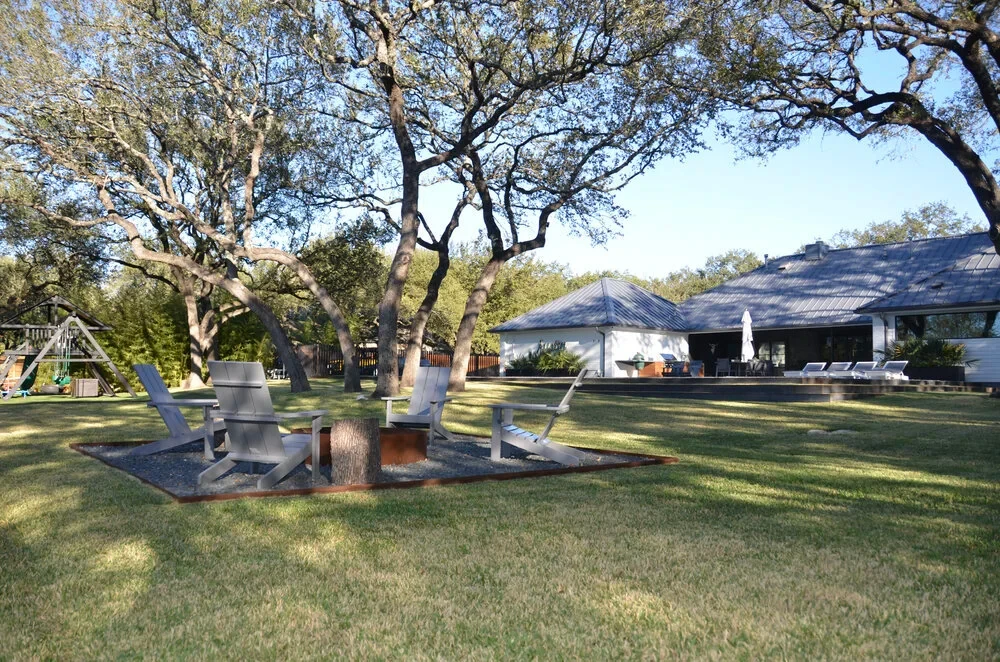 Chairs around a fire pit on an oak-shaded lawn facing a low white home in West Lake Hills