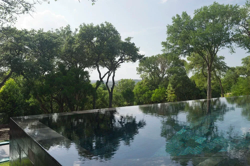 Infinity pool reflecting live oaks, its vanishing edge over the Hill Country treetops