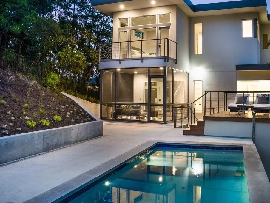Gunite pool with dark tile reflecting a lit modern home and balcony in Travis Heights at twilight