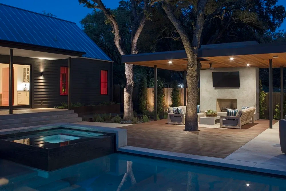 Pool and raised spa at twilight beside a covered patio with a fireplace, deck wrapping a live oak