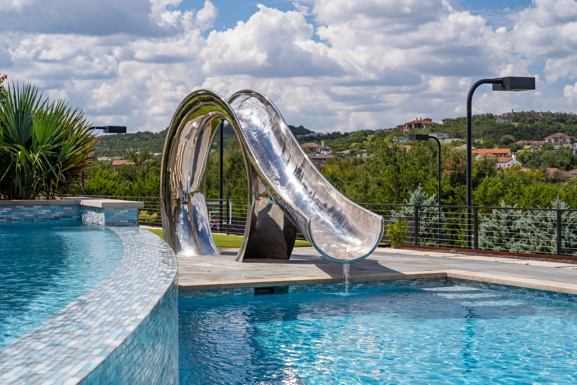 Polished stainless waterslide curving into a tiled pool, Lakeway hills and homes across the valley