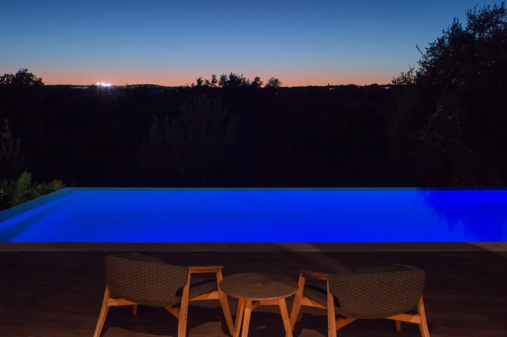 Infinity pool lit blue at dusk with lounge chairs, overlooking distant Hill Country lights