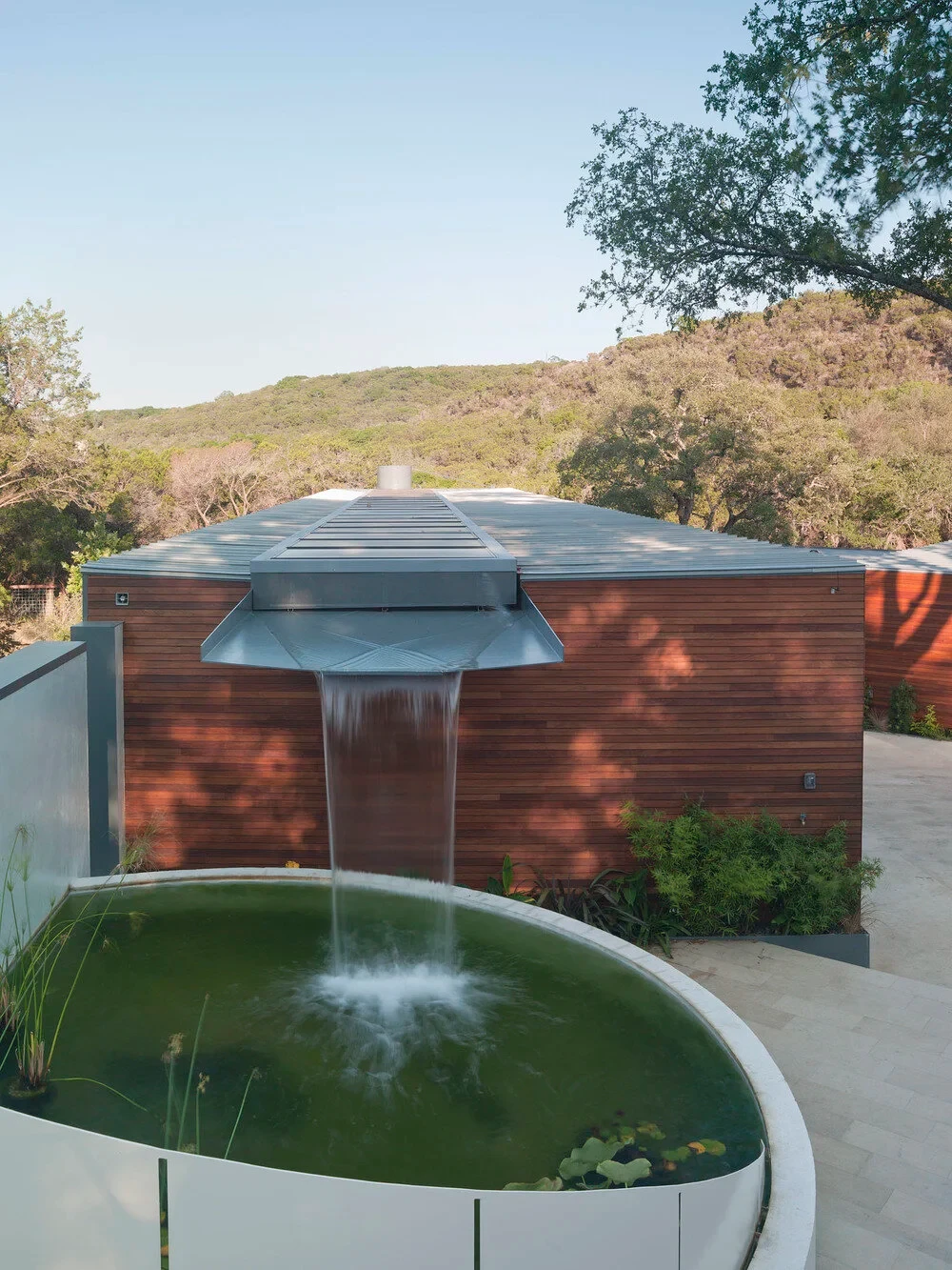 Sheer waterfall spilling off a wood-clad roof into a round stone pond pool, Hill Country beyond