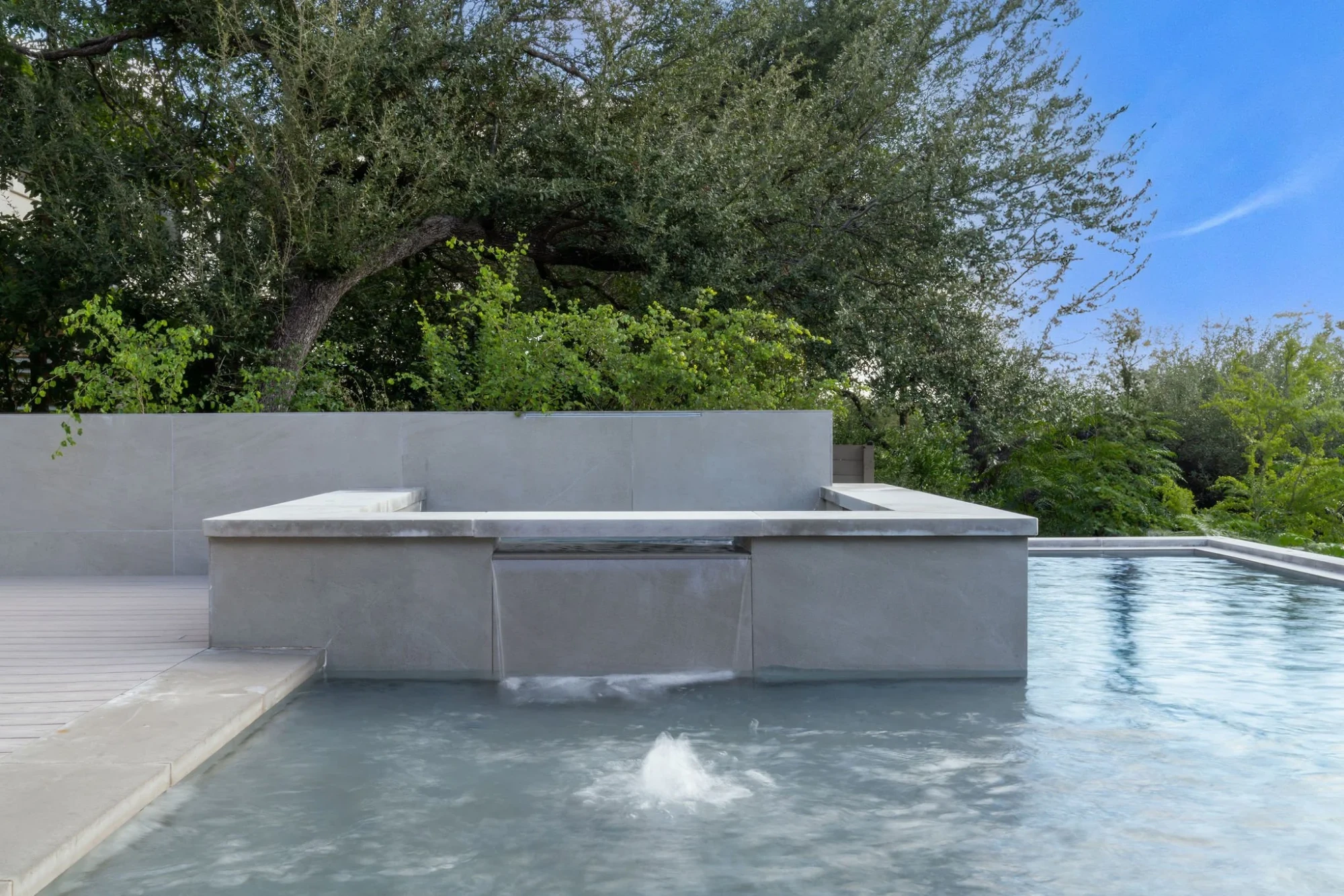 Concrete pool with a spillway from the raised spa, backed by a stone wall and an oak tree
