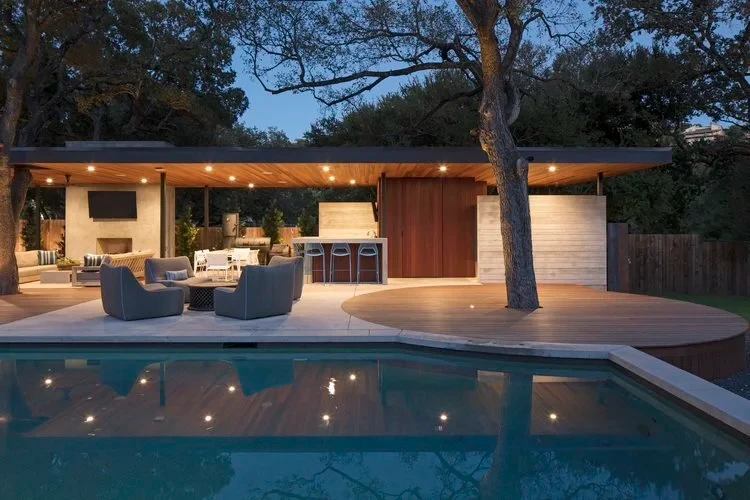Rectangular pool at dusk reflecting a lit pavilion with outdoor kitchen and lounge seating under oaks