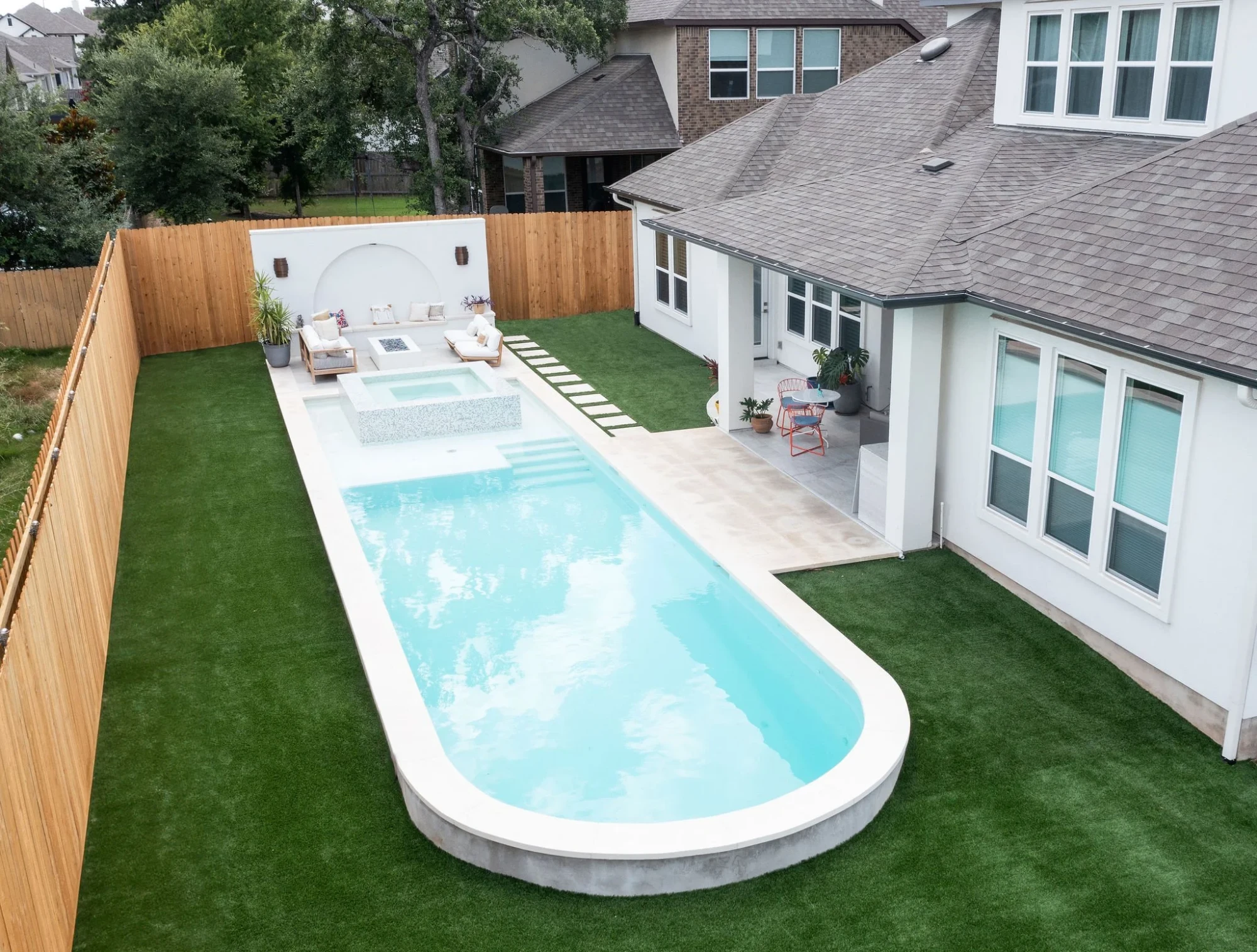 Aerial view of a long geometric pool with a raised spa, lounge seating and turf behind a two-story Austin home