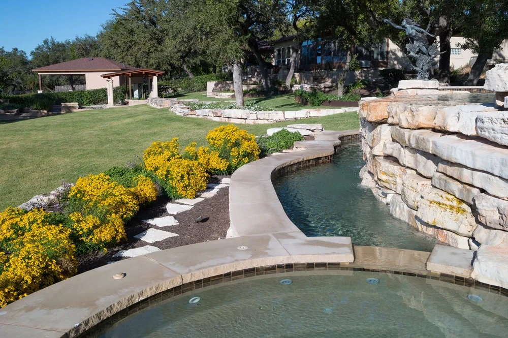 Stone-edged spa with native blooms and steps, a covered patio and lawn in the background