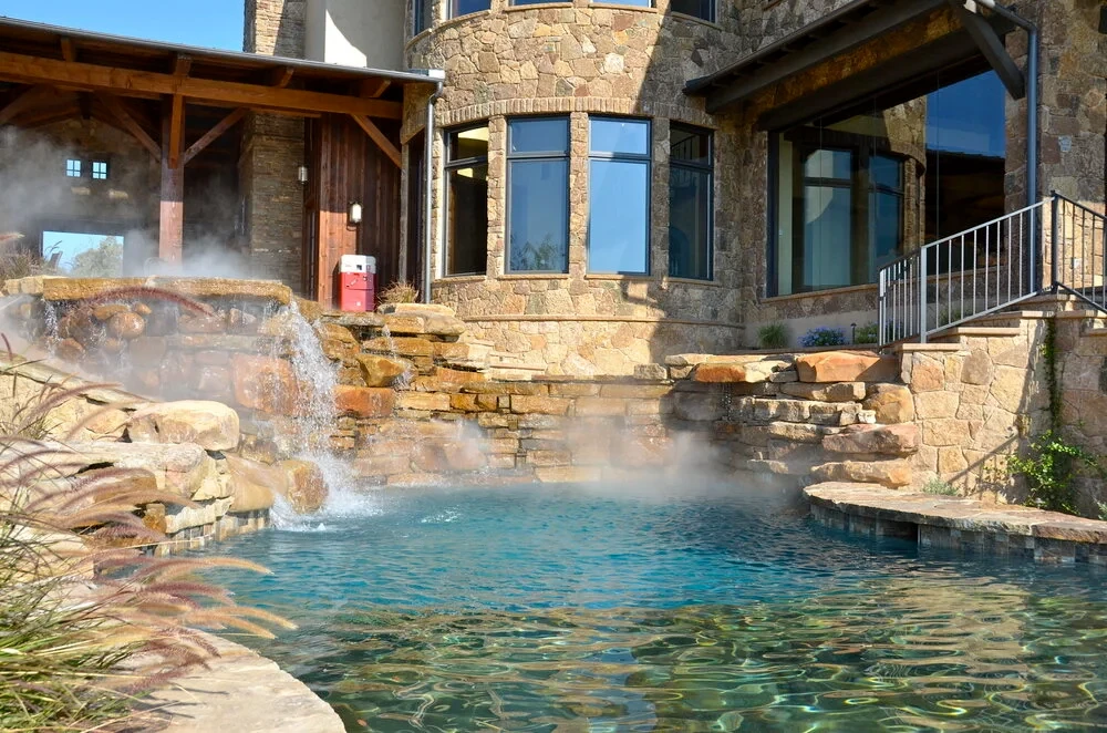 Steam rising off a freeform pool with stone waterfalls in a courtyard beside a stone Hill Country home