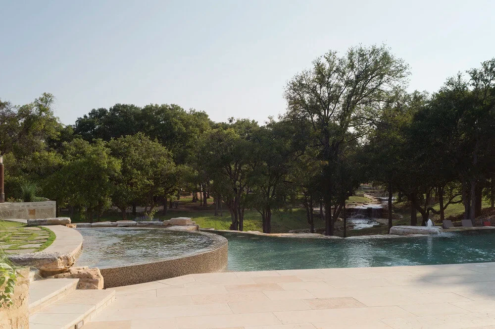Curving freeform pool edge and deck rolling into a grassy Hill Country lawn shaded by oaks