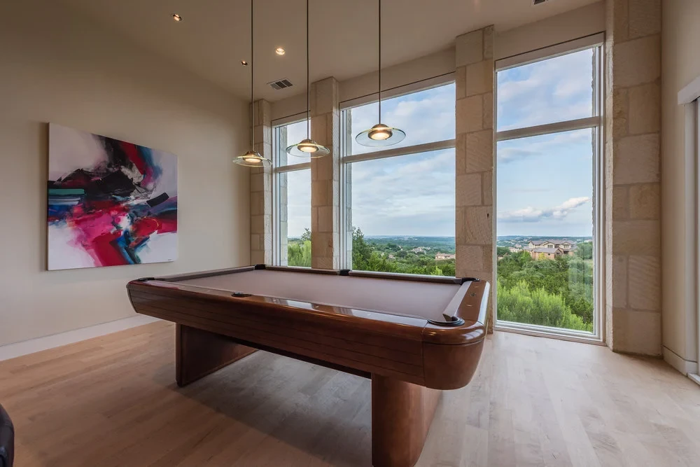 Game room with a billiard table and abstract art beside tall stone-framed windows over the Hill Country