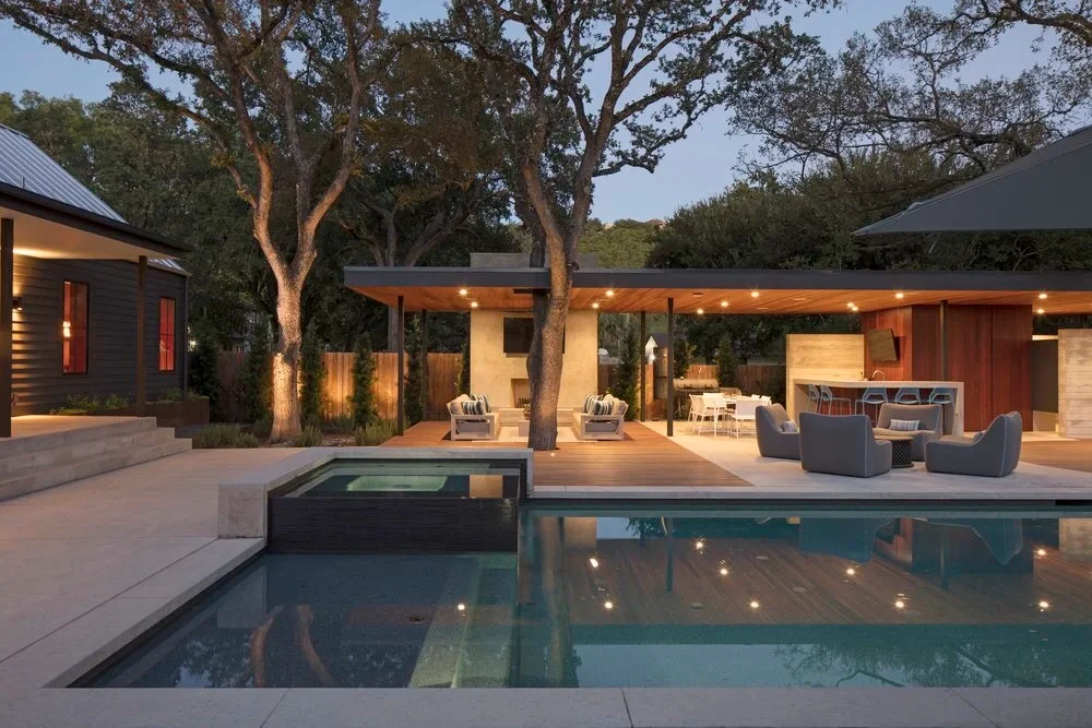Pool and spa glowing at twilight beside a covered patio with lounge seating under tall live oaks