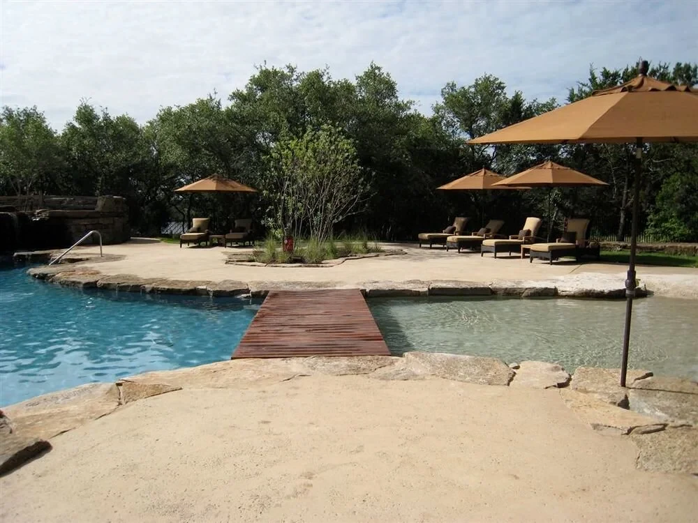 Freeform pool with a wood bridge, stone deck, and umbrellas over lounge chairs in Spanish Oaks