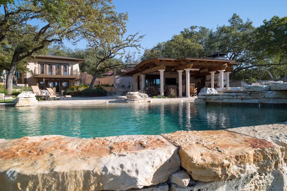 Freeform pool with a covered stone pavilion and outdoor kitchen, boulders along the flagstone deck