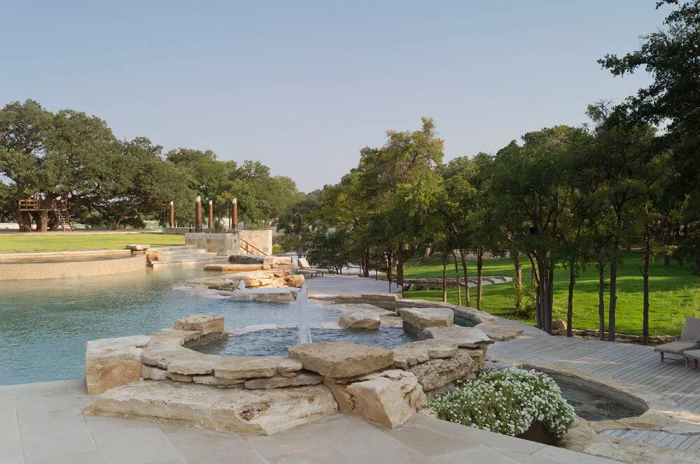 Freeform pool with a raised spa and stone deck on a lawn lined with oaks at dusk