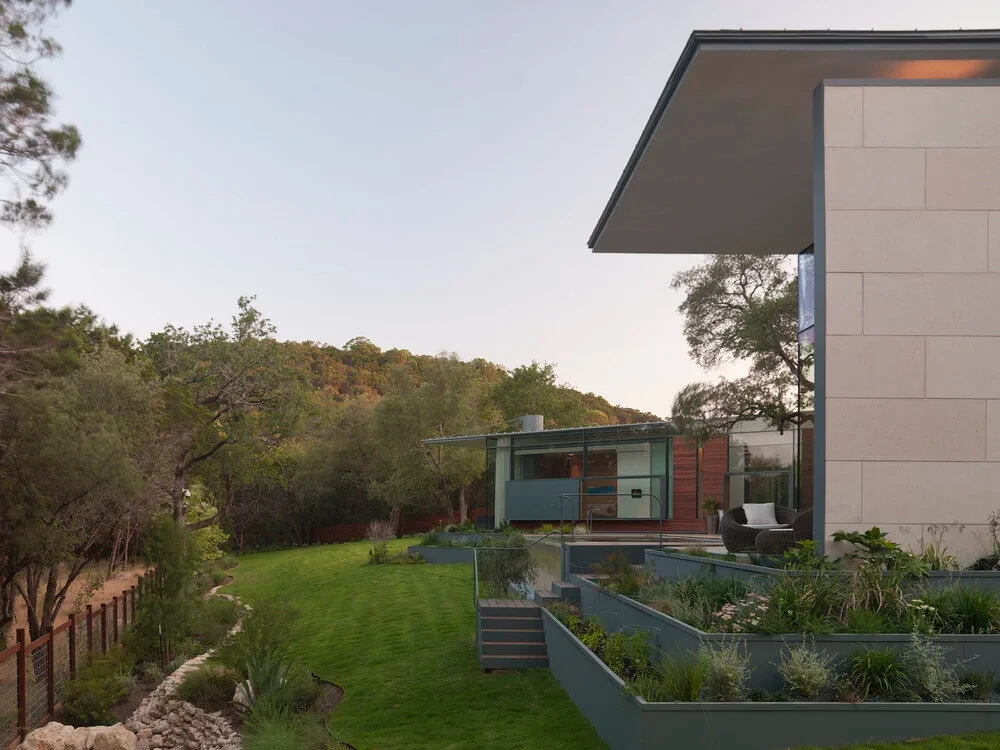 Stone modern home above a terraced lawn and planters, Hill Country ridgeline glowing at dusk