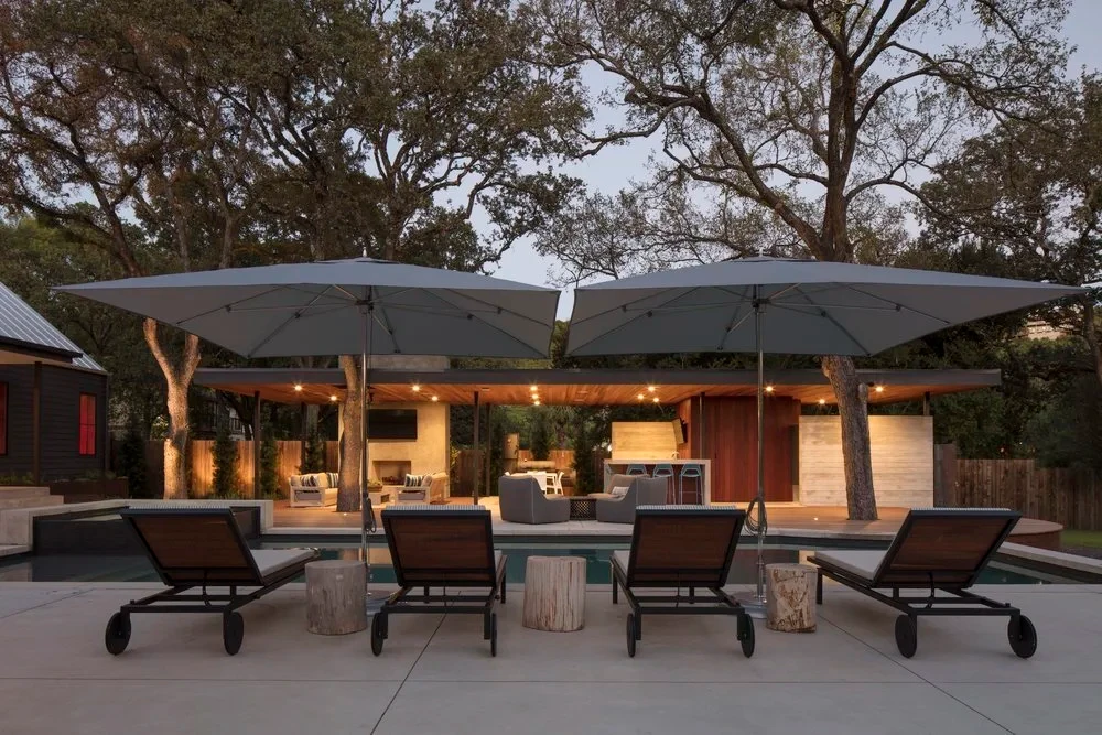 Cantilever umbrellas over lounge chairs on a patio at dusk, an outdoor kitchen lit beyond