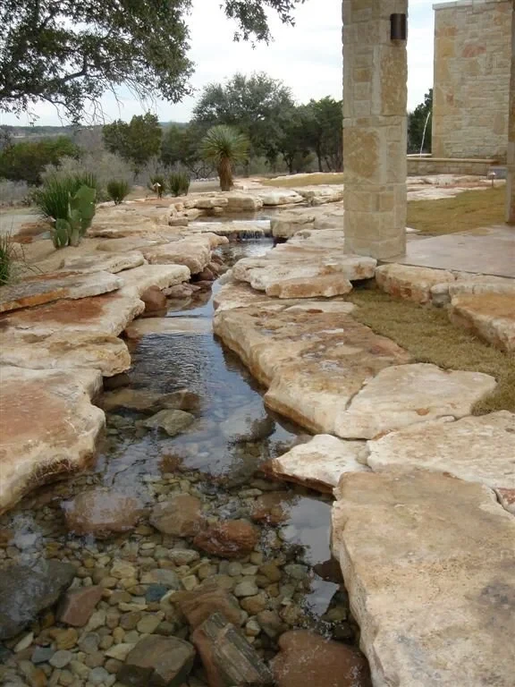 Stone-lined stream winding past limestone slabs and a column toward the Hill Country horizon