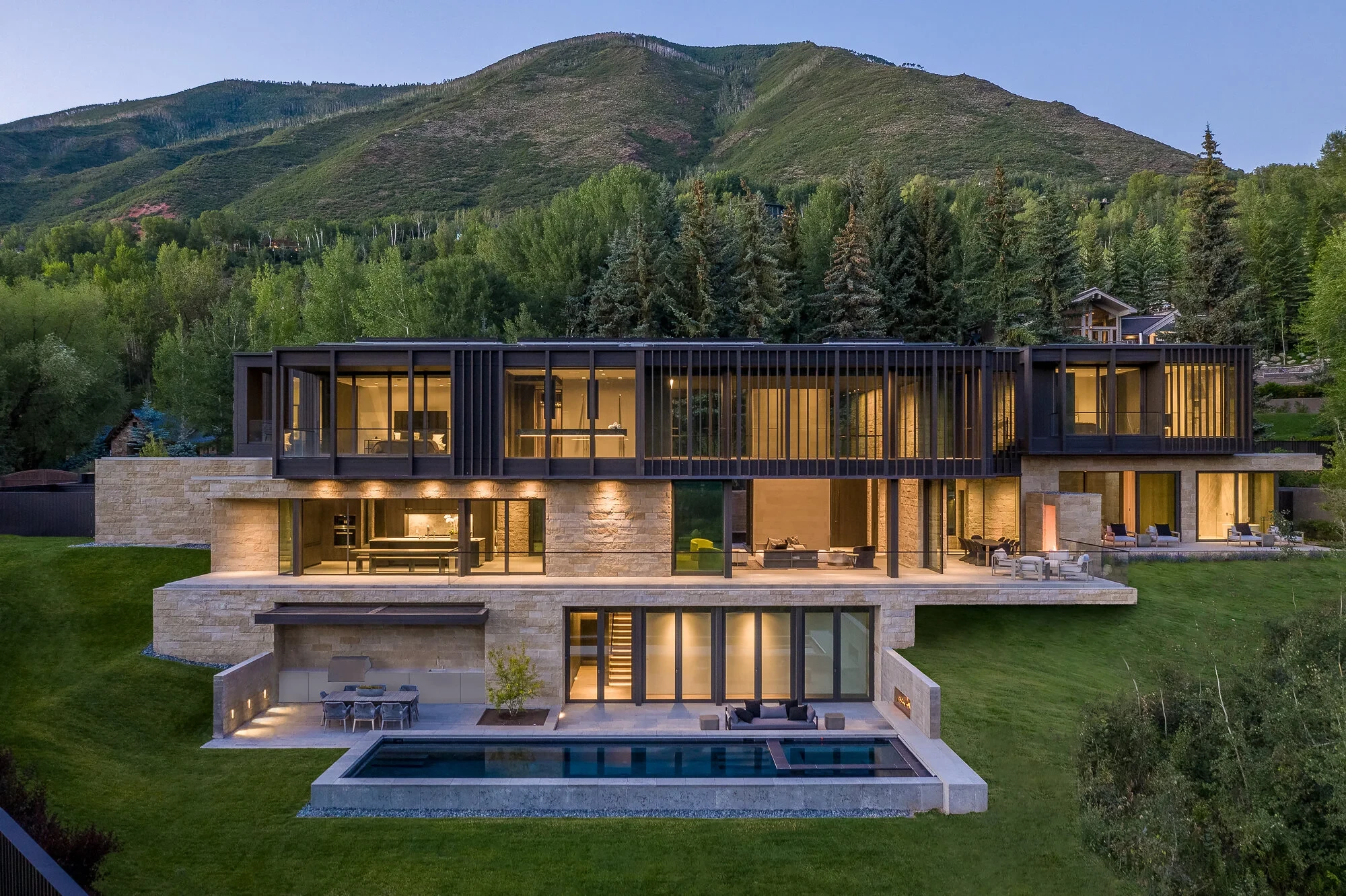 Modern stone home glowing at dusk with a geometric pool and lawn below a green Aspen mountainside