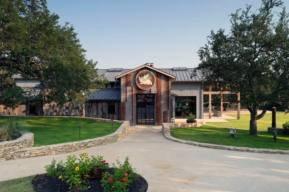 Timber-framed lodge with a metal roof and stone entry, framed by oaks on a landscaped drive