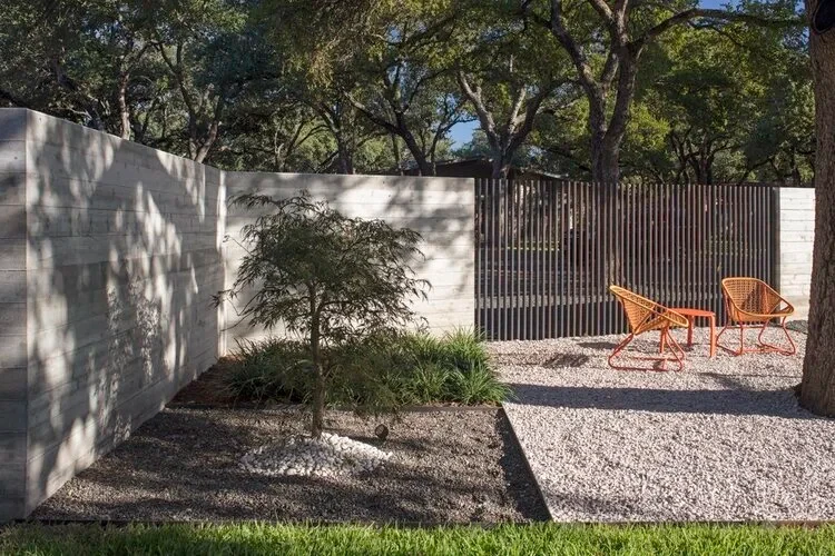 Concrete-wall courtyard with a small tree, gravel bed and orange wire chairs by a wood-slat gate